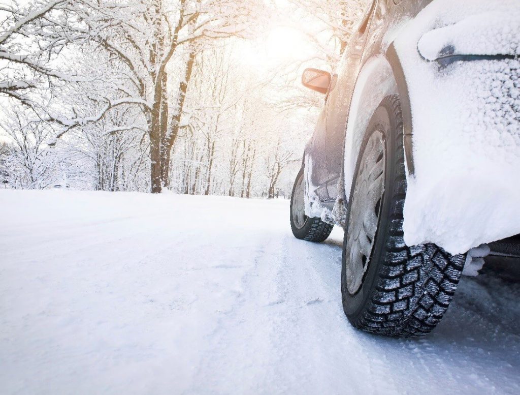 The side of a car driving on a wintery road. Surrounded by trees.