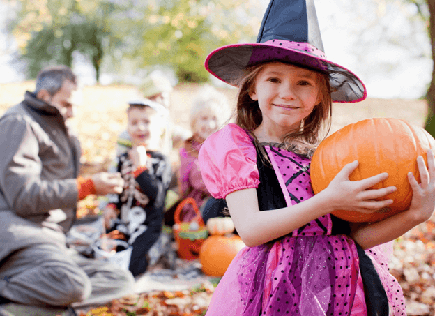 A family with pumpkins with the kids dressed up. The focus being a young girl as a witch holding a pumpkin.