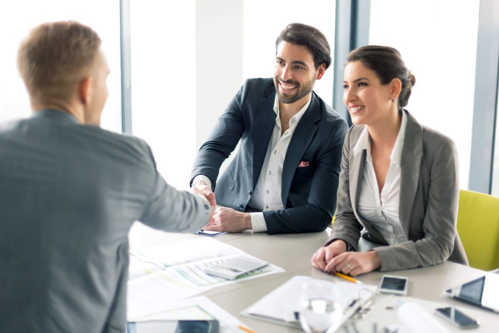 An insurance broker shaking hands with a client at a table with documents scattered across it.