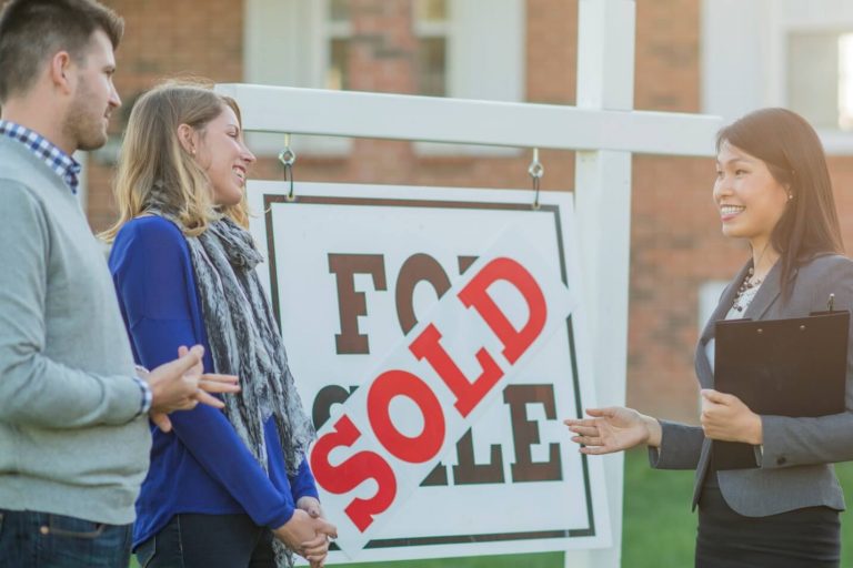 A realtor with a couple next to a for sale sign.