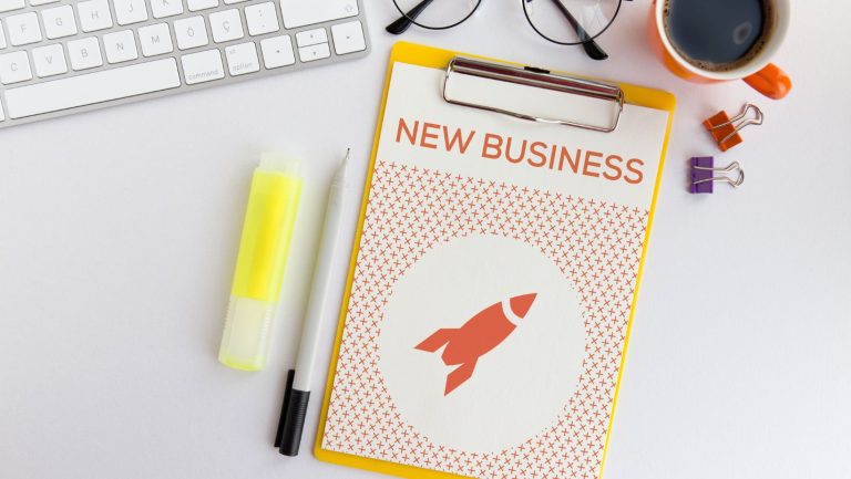 A clipboard with paper that says new business on a desk surrounded by different objects.