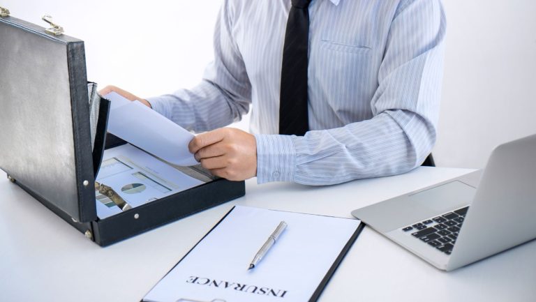 a business man sitting at a desk that has a open brief case, a laptop and a clipboard with paper that says insurance.