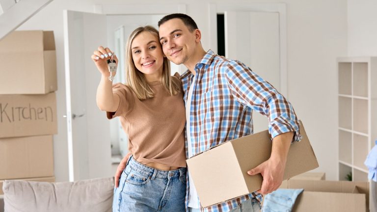a couple holding up their new home keys, surrounded by moving boxes.