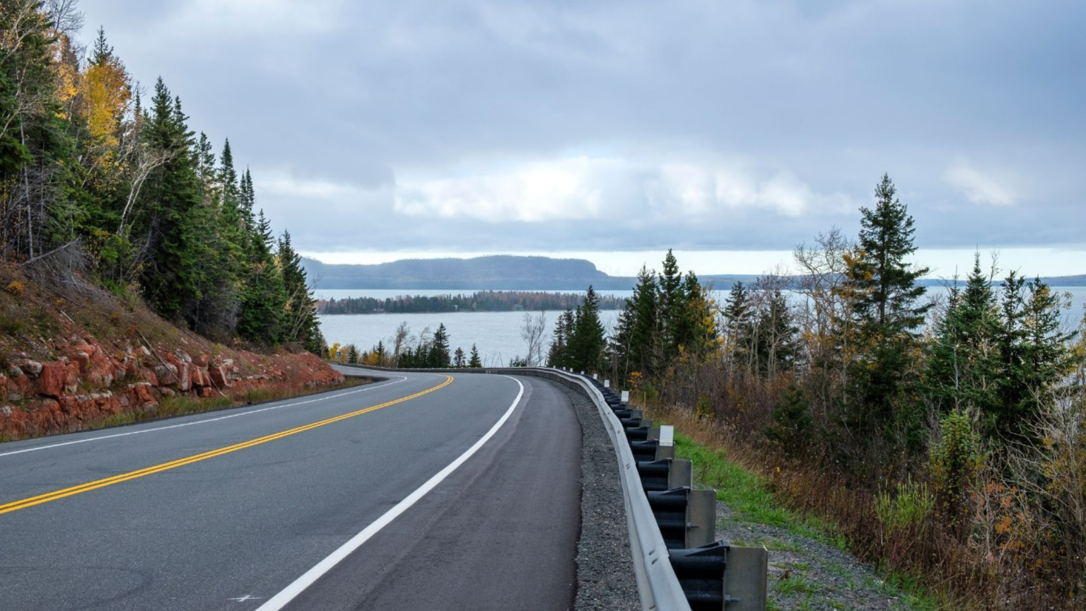 A winding road curves past dense evergreen trees and rocky terrain, overlooking a calm body of water under a cloudy sky. The scene is serene and expansive.
