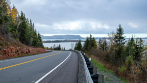 A winding road curves past dense evergreen trees and rocky terrain, overlooking a calm body of water under a cloudy sky. The scene is serene and expansive.