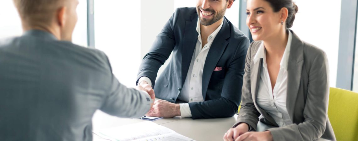 An insurance broker shaking hands with a client at a table with documents scattered across it.