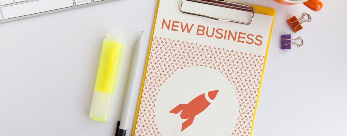 A clipboard with paper that says new business on a desk surrounded by different objects.