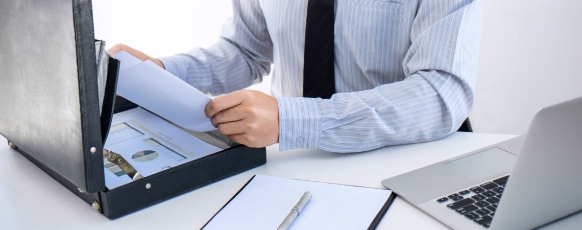 a business man sitting at a desk that has a open brief case, a laptop and a clipboard with paper that says insurance.
