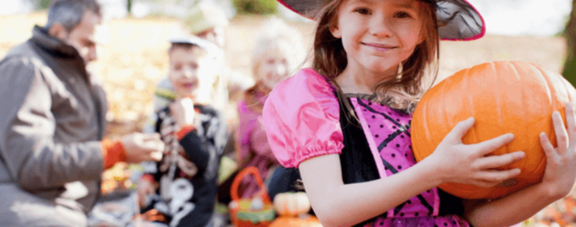 A family with pumpkins with the kids dressed up. The focus being a young girl as a witch holding a pumpkin.