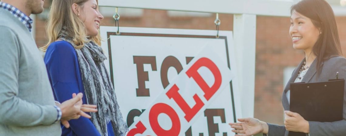 A realtor with a couple next to a for sale sign.